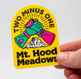 Hand holding a  yellow arch shaped sticker with 2 hands high five-ing. The text says, "Two Minus One" Across the arch and 
"Mt. Hood Meadows" at the bottom.