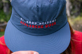 Blue cap with 'Mt. Hood Meadows Avalanche Dogs' text embroidered on it, held by a person wearing a red shirt.