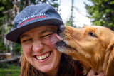 Person wearing a cap with 'Mt. Hood Meadows Avalanche Dogs' text, interacting with a dog outdoors.