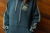A persons torso standing against a wooden wall, they are wearing a green hoodie with a pocket logo.