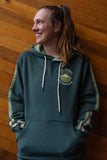 A person standing against a wooden wall wearing a green hoodie with a pocket logo.