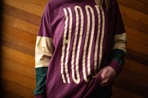 A persons torso against a wooden wall wearing a maroon and bone long sleeve shirt with 'HOOD' printed on it.