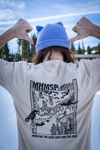 Person wearing a purple beanie and beige shirt with graphic design and text, outdoors.
