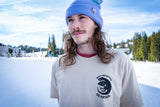 Person wearing a purple beanie and a beighe shirt with a logo, standing in a snowy landscape.